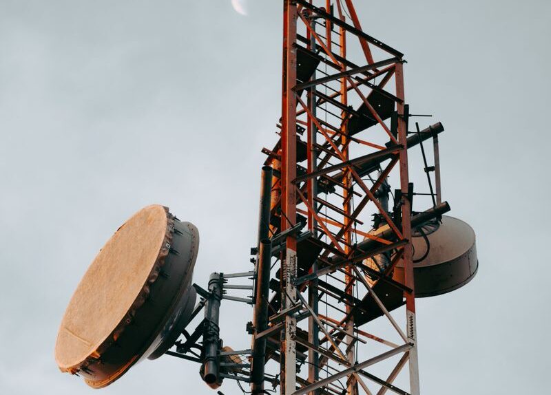 Vertical shot of a telecommunication tower with dishes under a cloudy sky and visible moon.