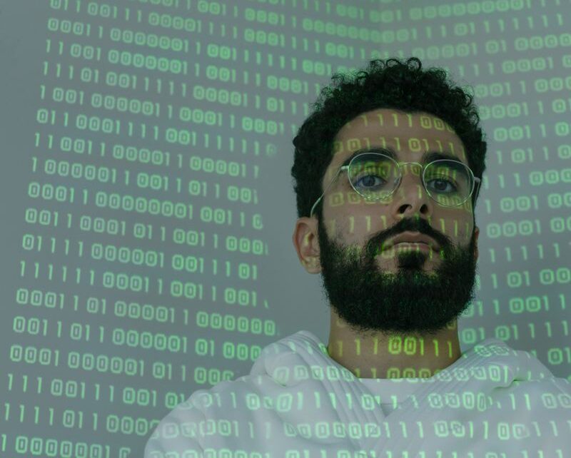 A bearded man in glasses stands with binary code projection, representing cybersecurity and digital intelligence.