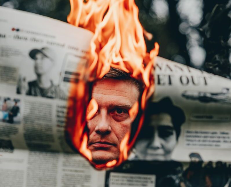 A man's face is revealed through a burning newspaper, creating an intense and striking visual.